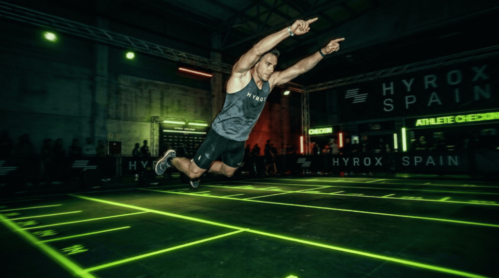 Atleta realizando el burpee broad jump en Hyrox con salto de longitud y estética neón industrial