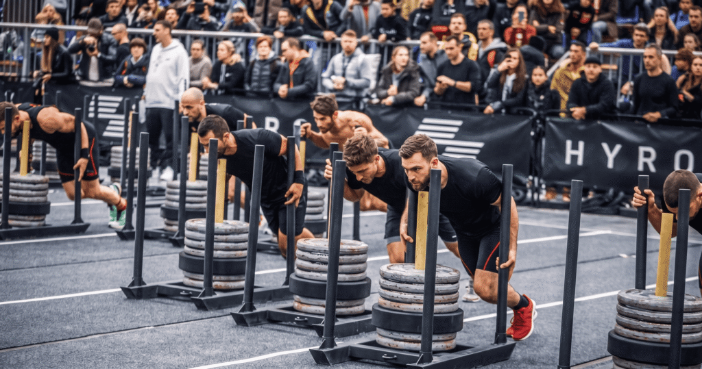 HYROX CATEGORIES & DIVISIONS: Wide-angle documentary shot in a large industrial arena showing four parallel hybrid athletes in synchronized sled push stances, demonstrating the standardized competitive environment across different race formats and class divisions.