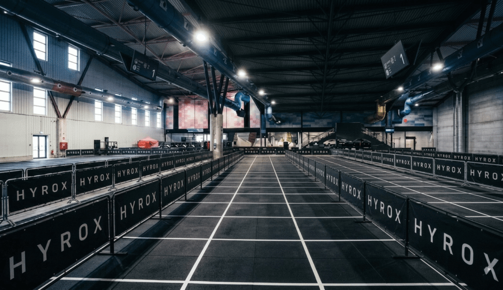 Perspective view of a standardized HYROX race floor grid with white-lined functional lanes and Roxzone transitions inside a professional indoor sports arena.