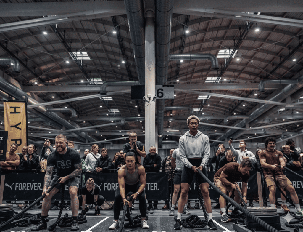 WHAT IS HYROX? Wide-angle documentary shot in a large industrial arena showing hybrid athletes pulling sleds in unison with a blurred crowd, capturing the standardized race atmosphere and global scale of the event.